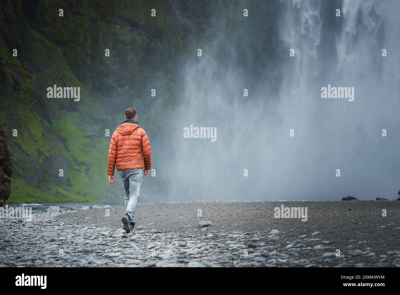 Persona in giacca arancione camminando verso la maestosa cascata Skogafoss in Islanda Foto Stock