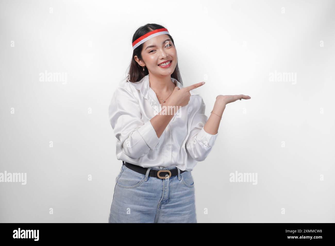 Donna asiatica eccitata che indossa un cerchietto con bandiera che presenta e punta lo spazio di copia sul lato, sorridendo ampio e in piedi su un backgroun bianco isolato Foto Stock
