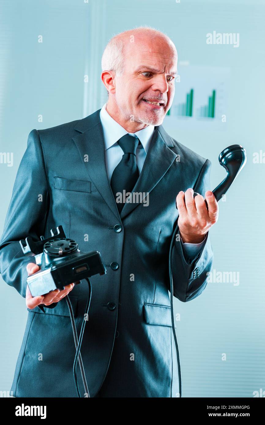 Un uomo d'affari furioso sta tenendo in mano un telefono fisso rotto, esprimendo la sua frustrazione per problemi di comunicazione o difficoltà tecniche in ufficio Foto Stock