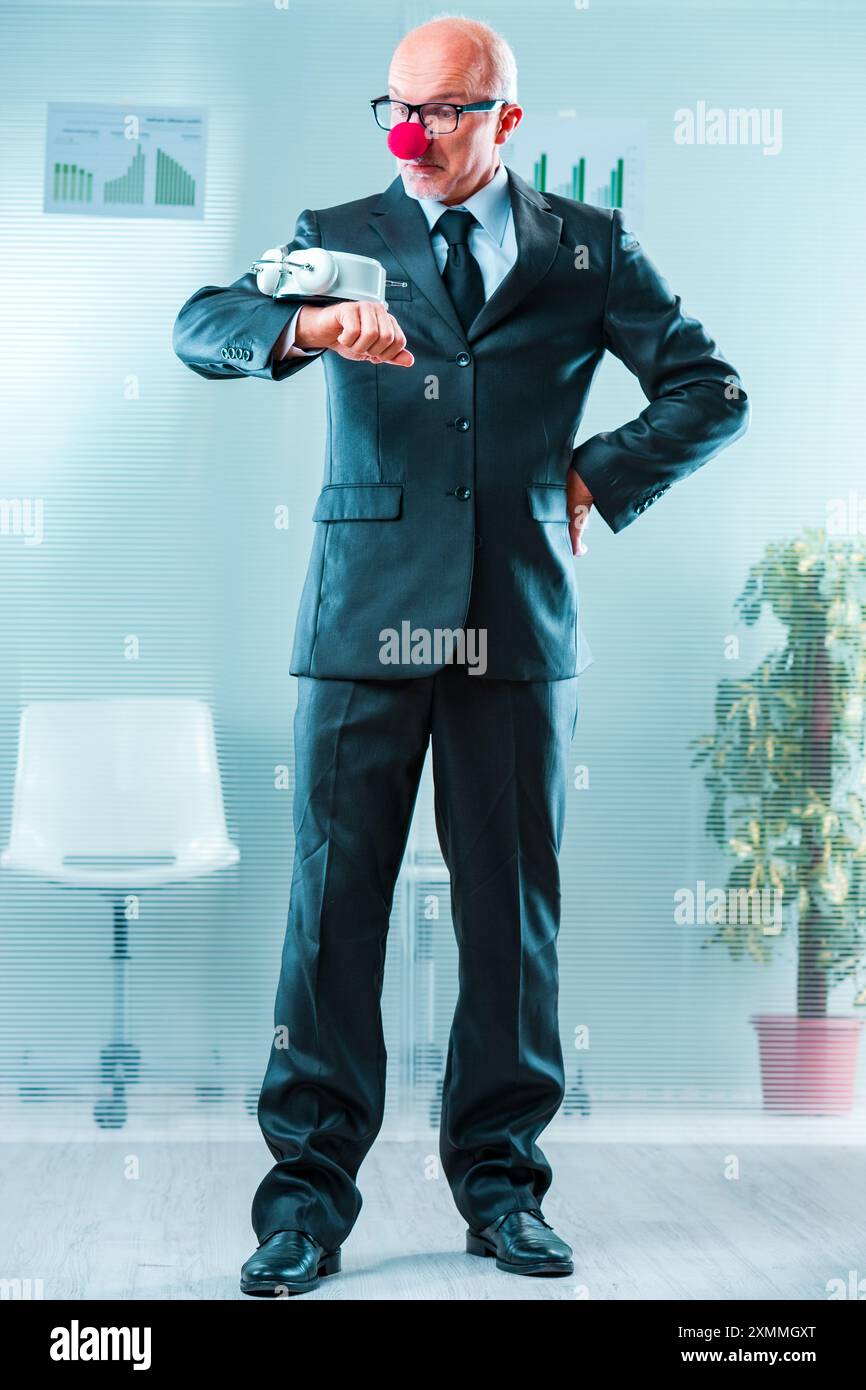 Un uomo dai capelli grigi in abito formale con il naso da clown regge un grande orologio, simboleggiando la ridicolezza delle scadenze pressanti a causa della cattiva gestione. B Foto Stock