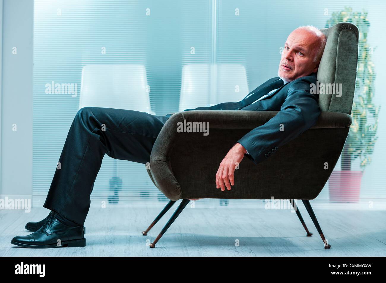 L'anziano uomo d'affari in costume si aggrappa su una sedia, sembra esausto e disperato, temendo che l'intelligenza artificiale sostituisca i posti di lavoro. Sfondo sfocato per ufficio Foto Stock