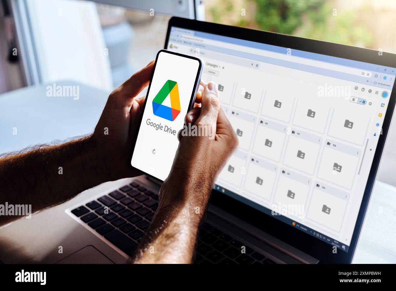 Google Drive sul telefono cellulare e sul computer. Uomo che possiede uno smartphone con un'app di archiviazione cloud sullo schermo. Laptop in background che mostra le cartelle google Foto Stock