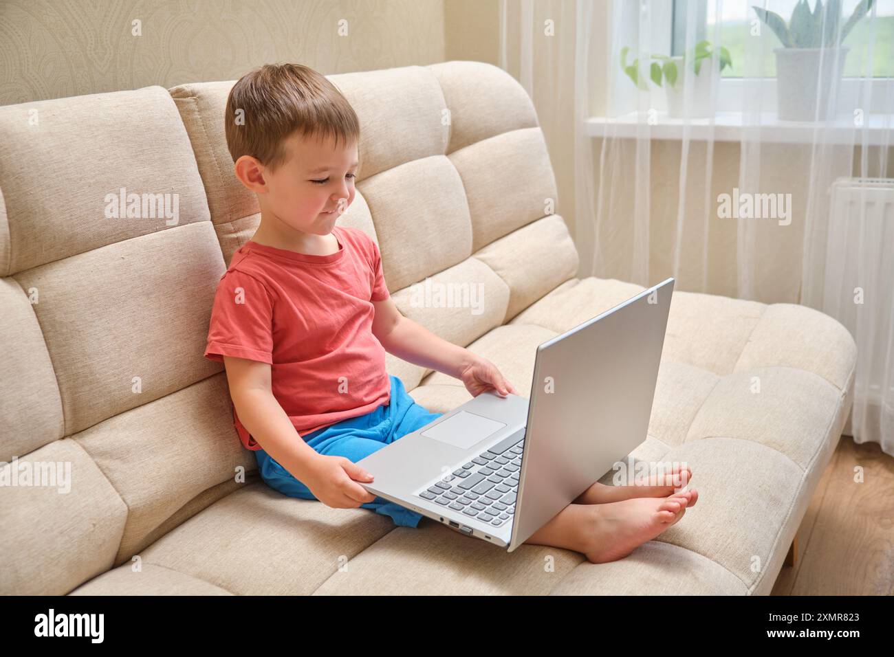 Un ragazzo è seduto su un divano con un portatile in grembo. Indossa una camicia rossa e pantaloncini blu. Bambino di 3 anni (bambino di 3 anni) Foto Stock