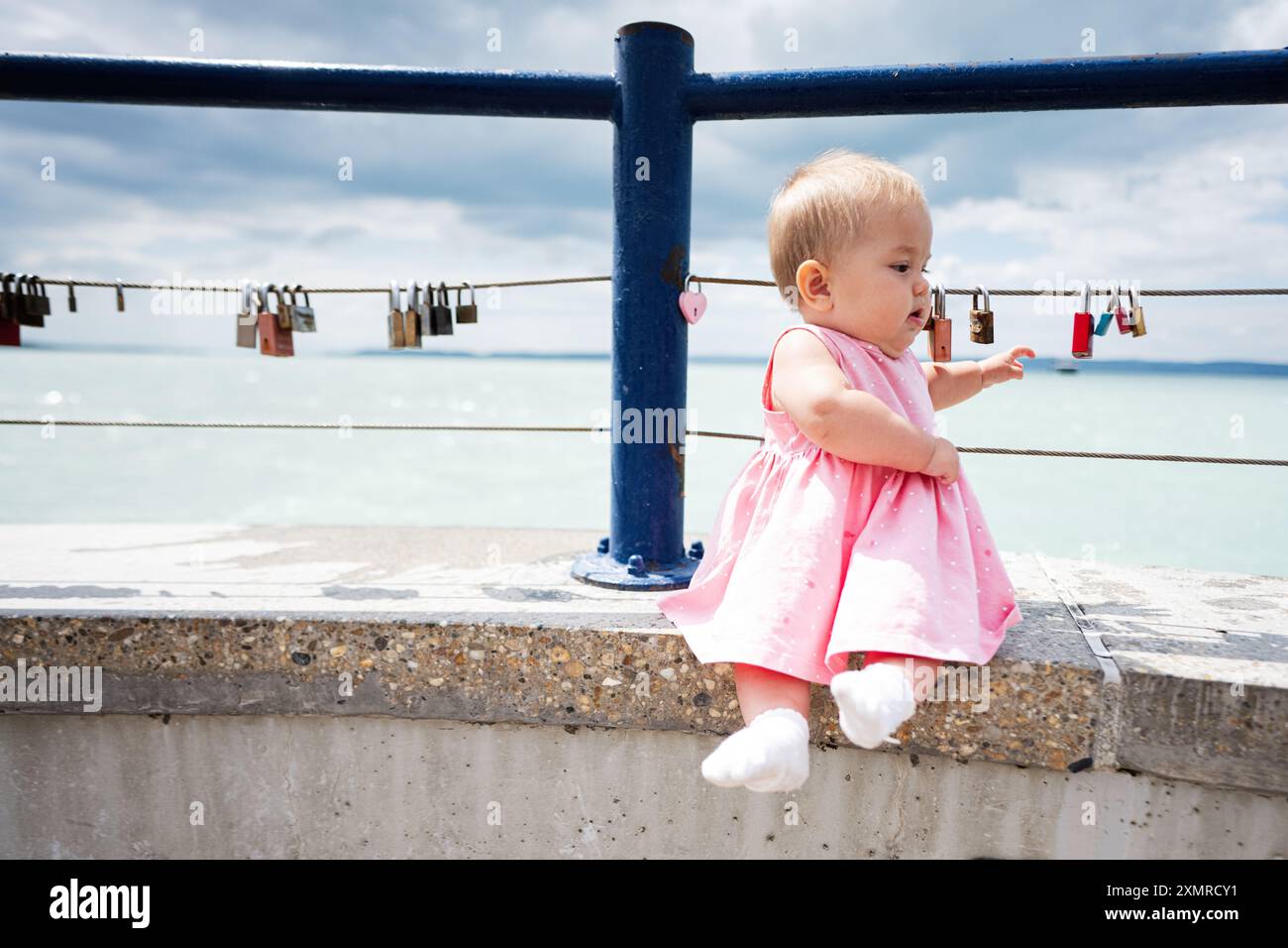 Adorabile bambina in abito rosa seduta accanto al mare. Vista sul mare con lucchetti sulle ringhiere. Momenti indimenticabili catturati all'aperto. Foto Stock