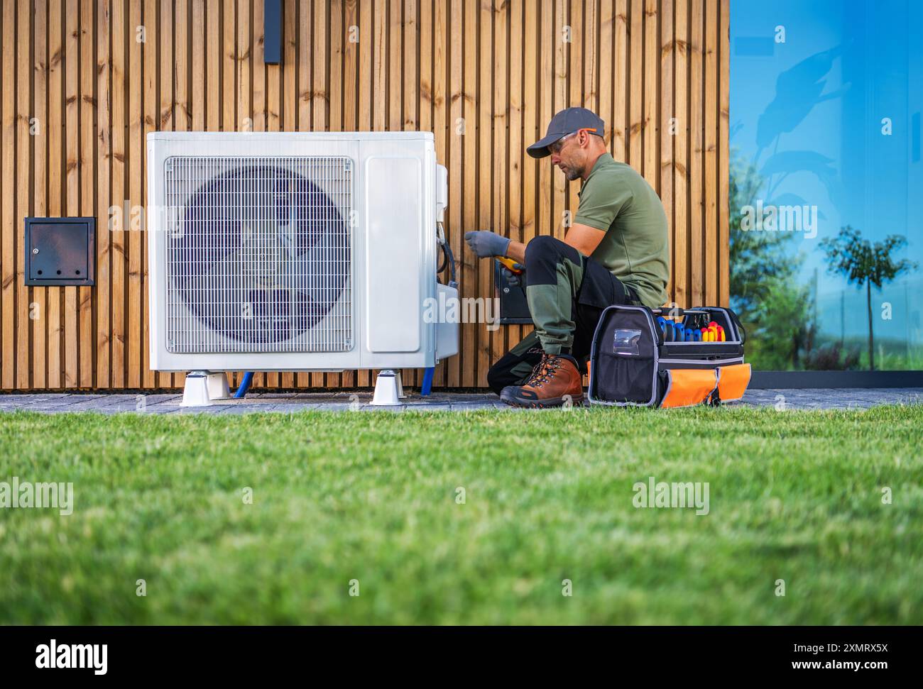 Un tecnico sta effettuando la manutenzione di un'unità di aria condizionata situata accanto a una casa moderna. Indossando abiti da lavoro, utilizza gli attrezzi di una cassetta degli attrezzi organizzata Foto Stock