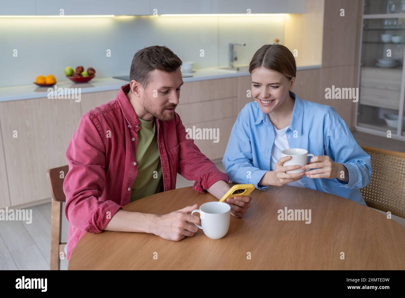 Uomo contento che si mostra a una donna smartphone a casa. Coppia felice seduta con tazze al tavolo della cucina Foto Stock