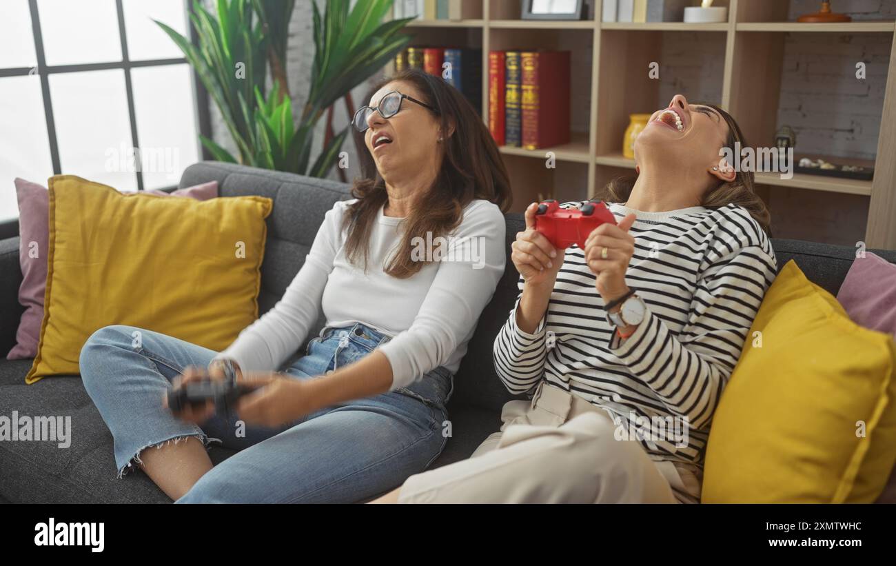 Due donne ridono gioiosamente su un divano tenendo in mano i controller di gioco in un accogliente salotto. Foto Stock