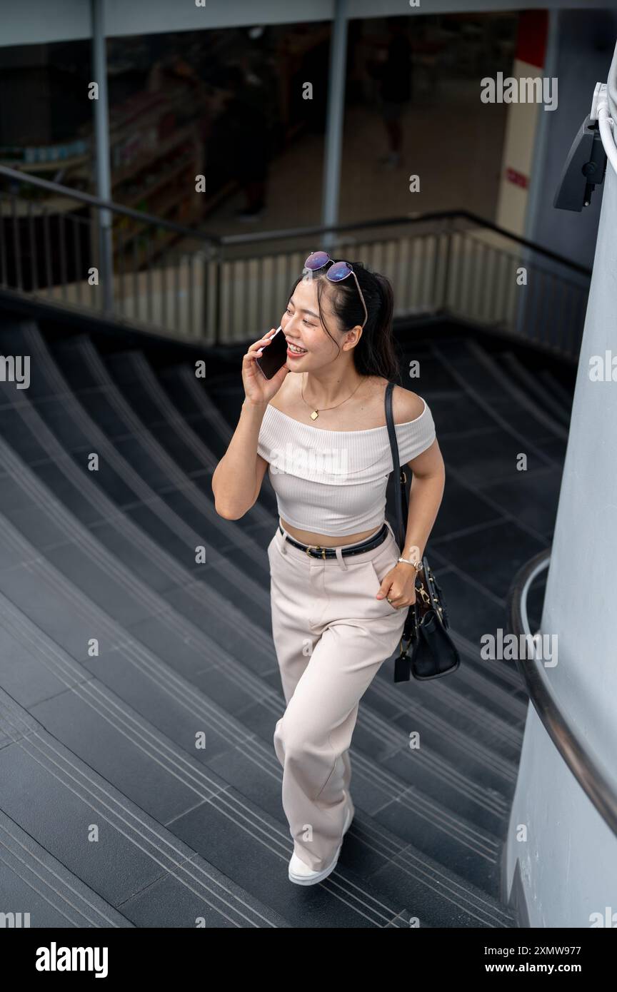 Una bella donna asiatica in un abito alla moda parla al telefono mentre sale le scale in città. stili di vita, vita in città, tecnologia wireless Foto Stock