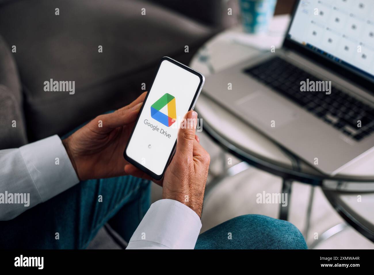 Google Drive sul telefono cellulare e sul computer. Uomo che possiede uno smartphone con un'app di archiviazione cloud sullo schermo. Laptop sullo sfondo che mostra l'unità google Foto Stock
