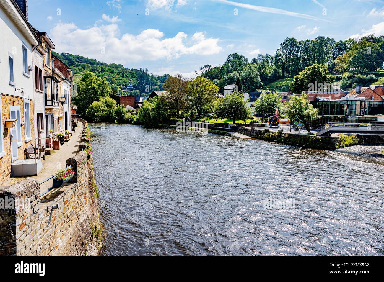 Fiume Ourthe nel villaggio di la Roche-en-Ardenne, muri di pietra, case, edifici e percorso pedonale lungo la riva del fiume, montagne con alberi verdi in mis Foto Stock