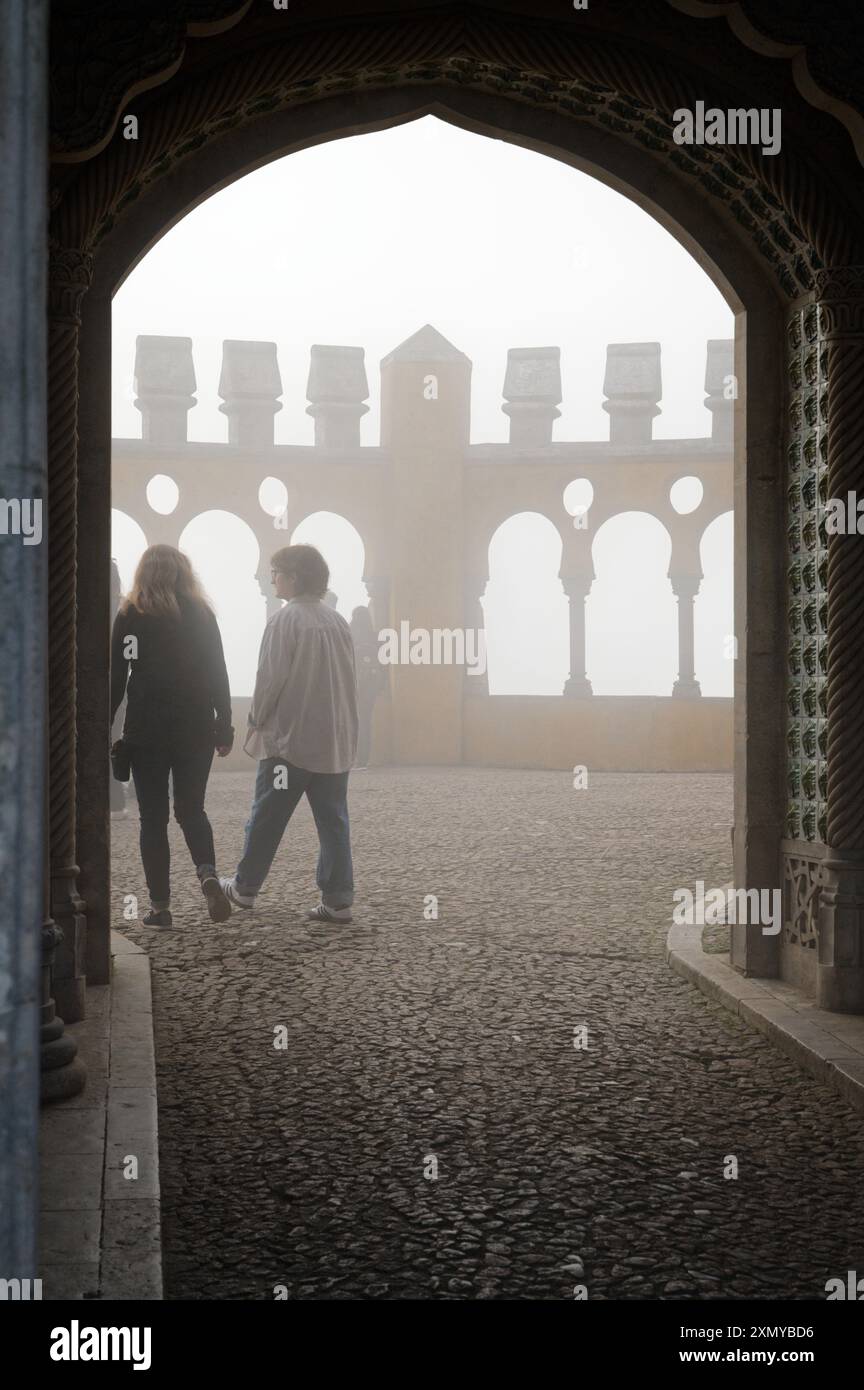 Due visitatori passeggiano attraverso un arco nebbioso a Palazzo pena, incorniciato da archi gotici e immerso nella luce soffusa ed eterea di una giornata nebbiosa Foto Stock