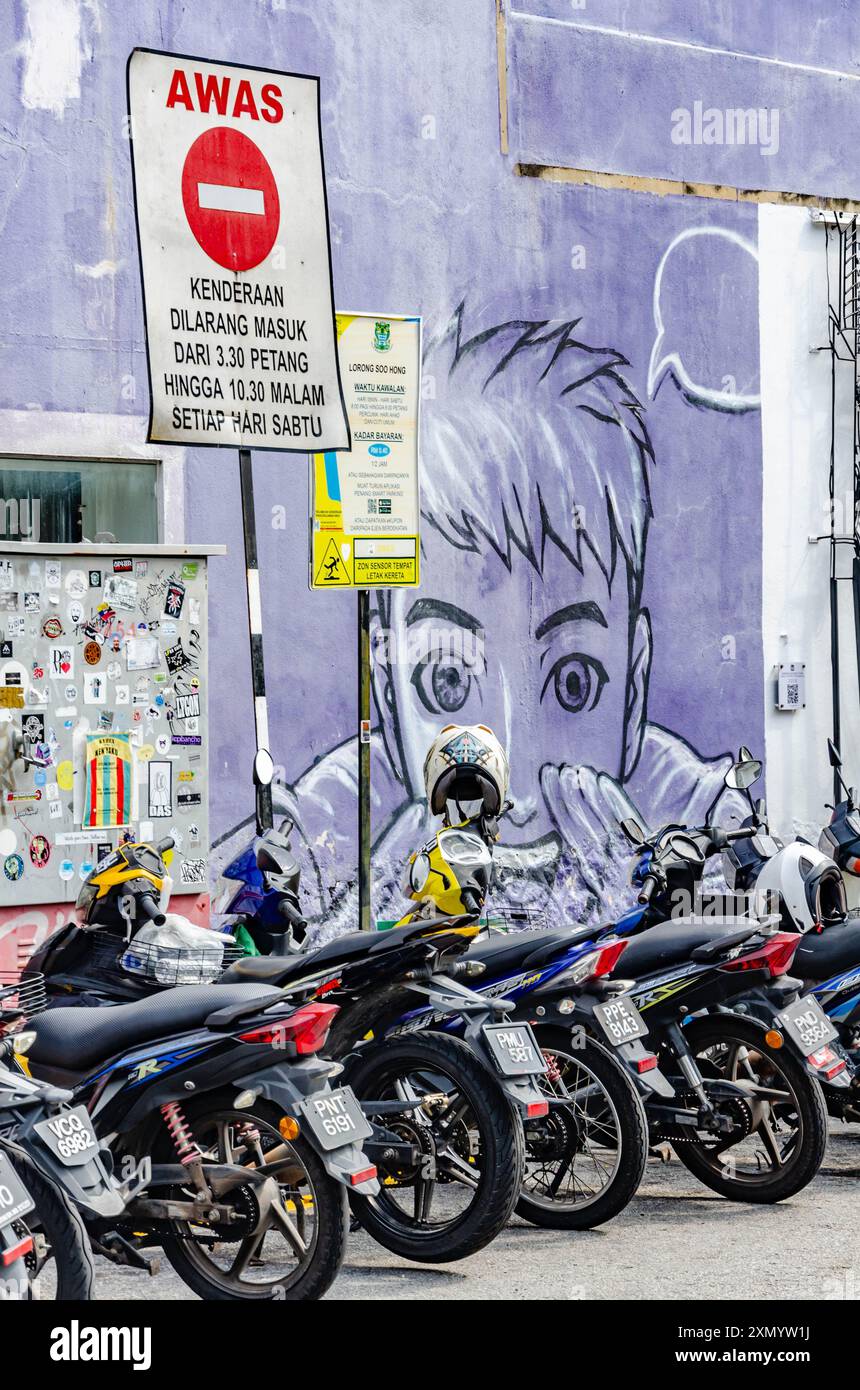 Una fila di motociclette parcheggiate sul lato di una strada di fronte a un muro viola con un grande murale in Armenian Street a George Town, Penang, Malesia Foto Stock