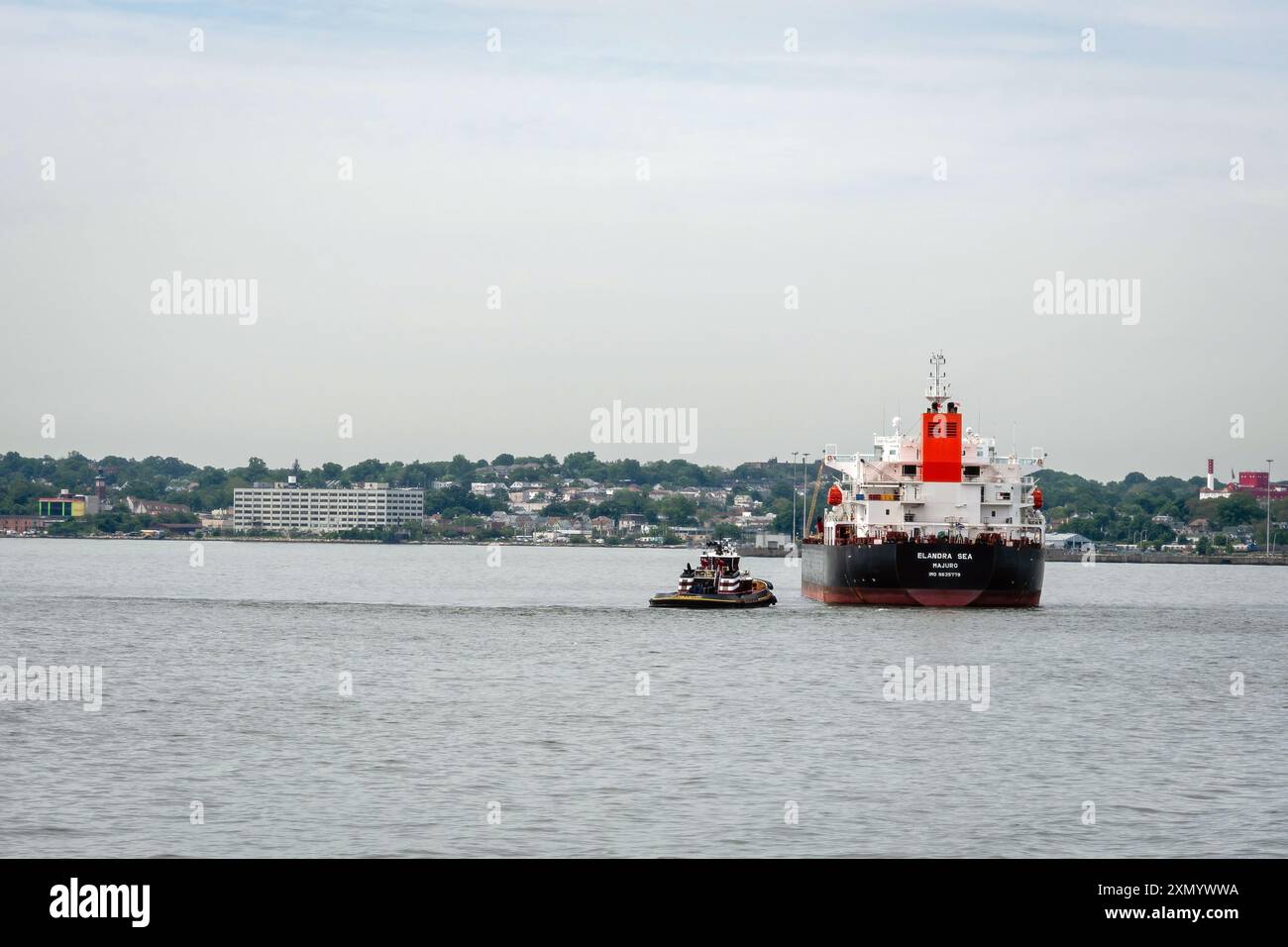 New York, USA - 7 giugno 2019: Un rimorchiatore guida una nave da carico attraverso le acque calme vicino alla città. Foto Stock