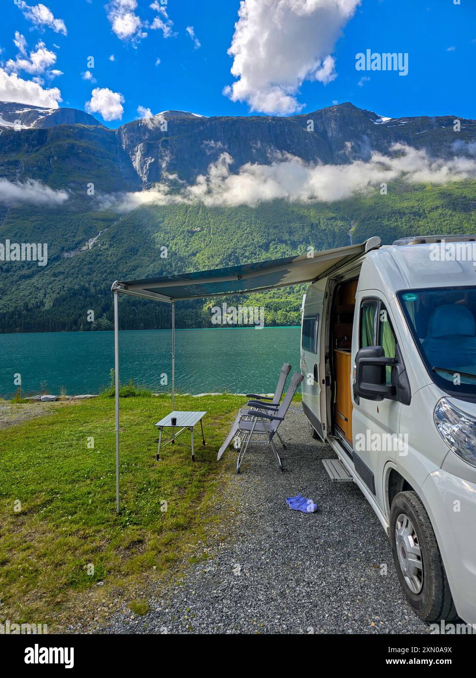 Un accogliente camper si trova accanto alle acque tranquille, incorniciato da torreggianti montagne e lussureggiante vegetazione sotto un cielo vivido. Lovatnet, Norvegia Foto Stock
