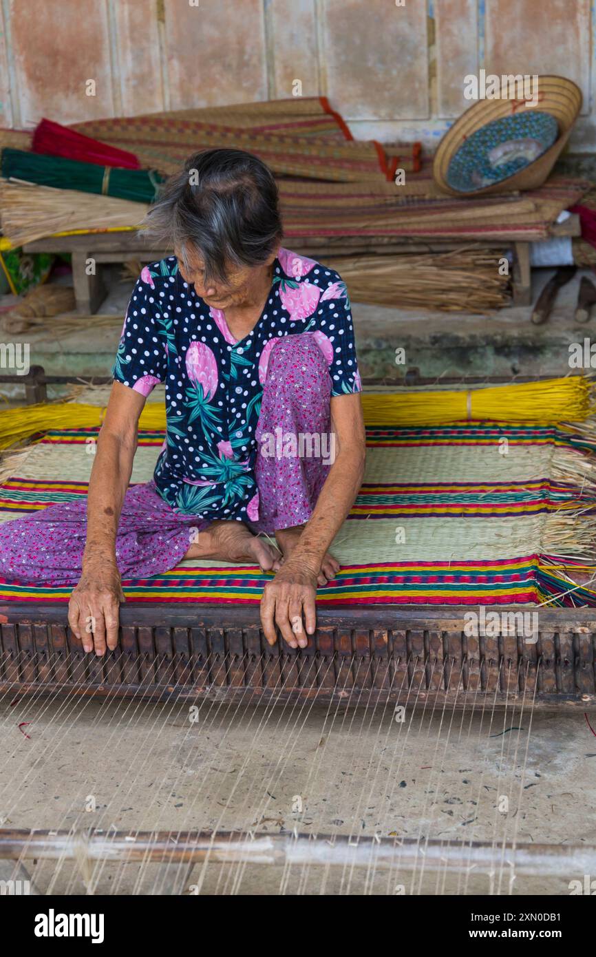 Anziana donna vietnamita che tesseva materassini da canne essiccate e tinte su telaio nel villaggio di tra Nhieu, Quảng Nam, Vietnam centrale, Asia a giugno Foto Stock