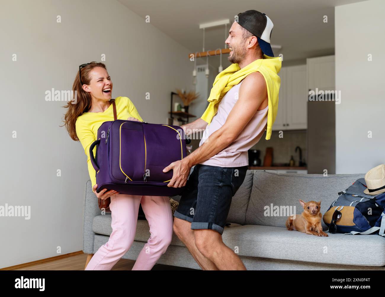 Trasloco a casa o preparazione per una vacanza estiva. Felice giovane coppia solleva una borsa da viaggio e ridendo, un piccolo cane giace sul divano. Foto Stock