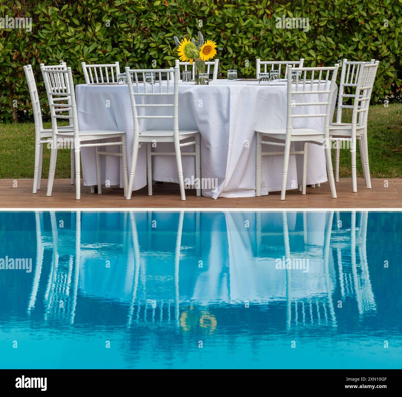 Tavolo preparato per cena, con sedie bianche di tovaglia e vaso con girasoli gialli, che si riflette sull'acqua blu della piscina Foto Stock