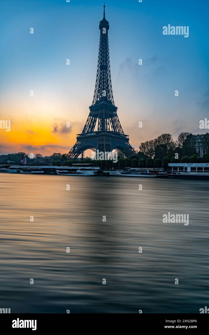 La Torre Eiffel si erge maestosamente contro un colorato cielo mattutino mentre il sole sorge, gettando riflessi sulle calme acque della Senna nelle vicinanze. Foto Stock