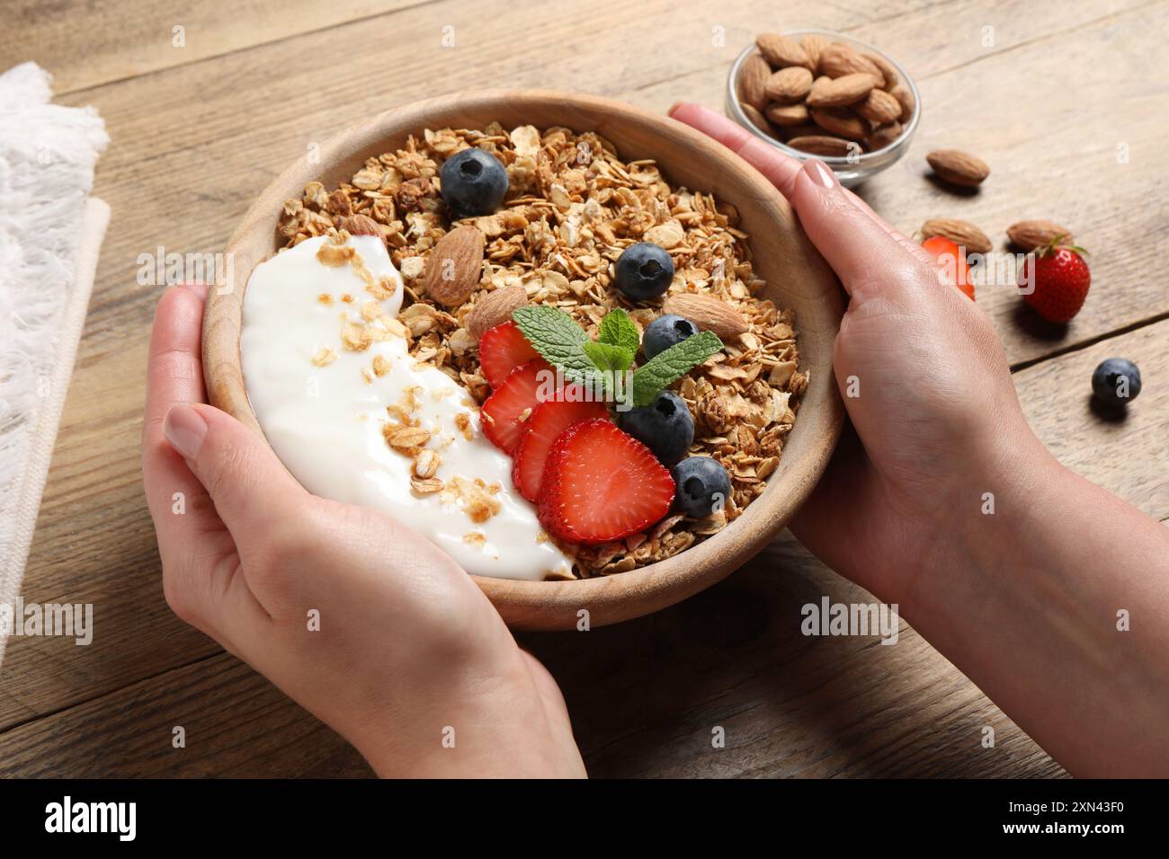 Donna che tiene una ciotola di gustose granole con frutti di bosco, mandorle e yogurt al tavolo di legno, primo piano Foto Stock