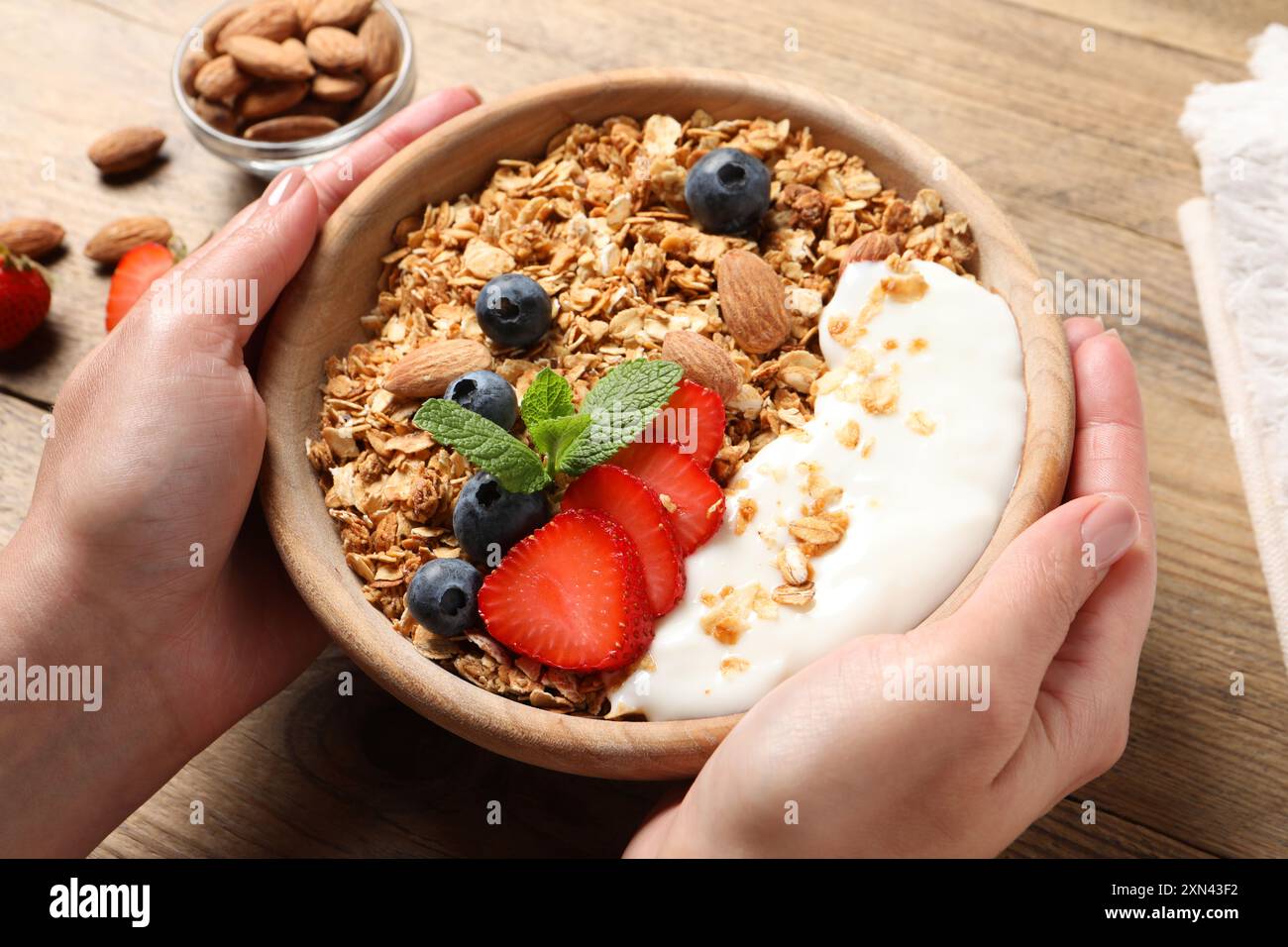 Donna che tiene una ciotola di gustose granole con frutti di bosco, mandorle e yogurt al tavolo di legno, primo piano Foto Stock