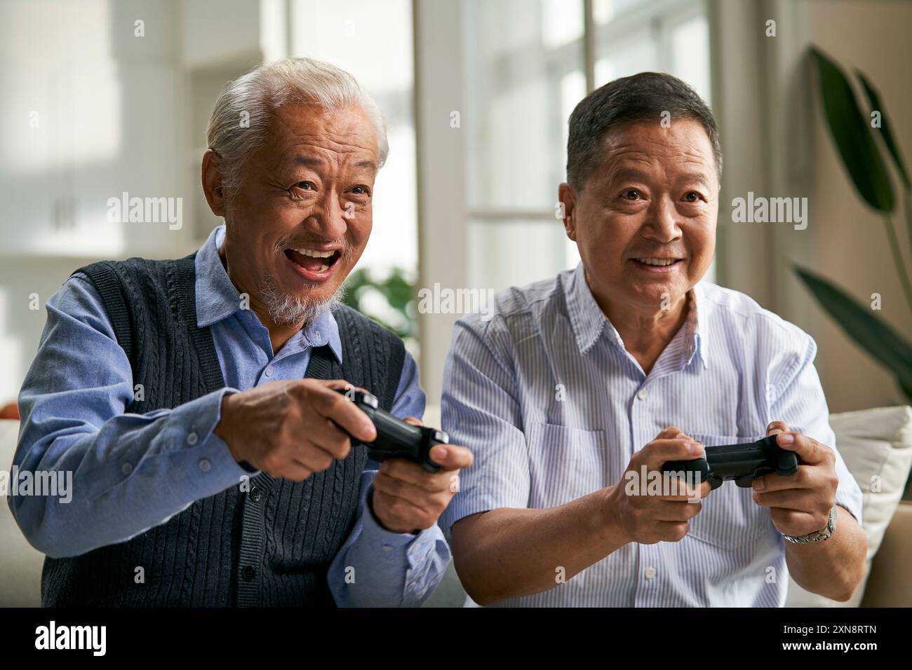 due uomini asiatici anziani felici seduti sul divano a casa a giocare a videogame Foto Stock