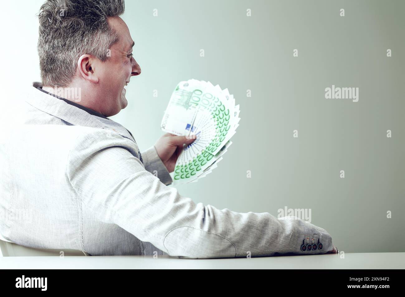 Un uomo d'affari è seduto alla sua scrivania, ridendo e tenendo in mano un fan delle banconote in euro, che rappresenta la sua avidità e le sue pratiche commerciali non etiche Foto Stock