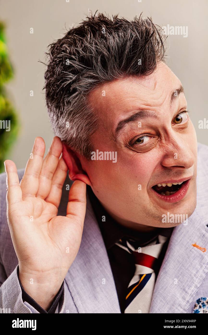 Un uomo d'affari con un'espressione astuta sta ascoltando intensamente, tenendo la mano stretta all'orecchio, cercando di ascoltare una conversazione privata Foto Stock