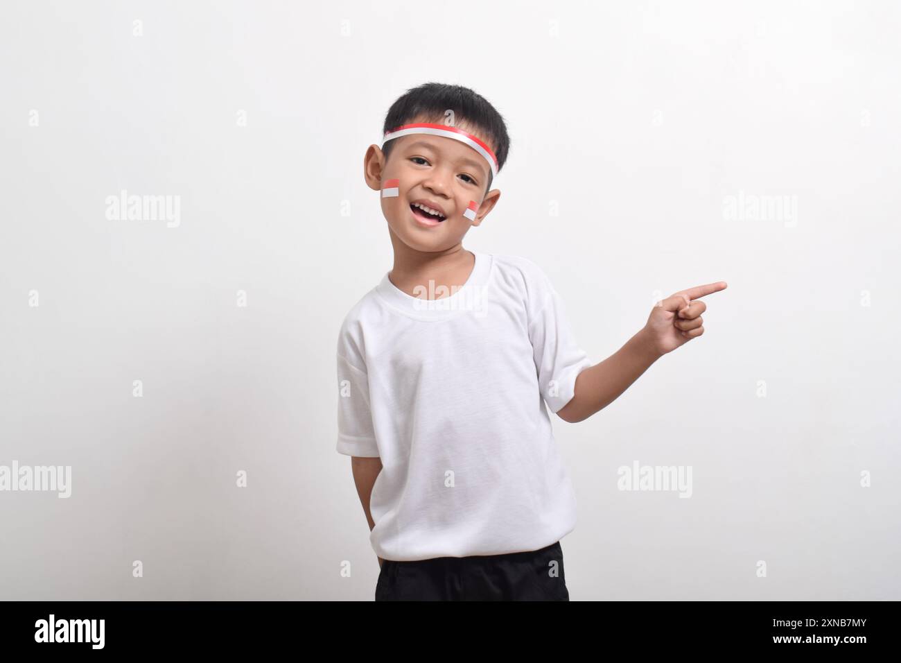 Ragazzo asiatico che indossa un cerchietto con bandiera che punta allo spazio di copia accanto a lui isolato su sfondo bianco. Concetto della giornata dell'indipendenza dell'Indonesia Foto Stock