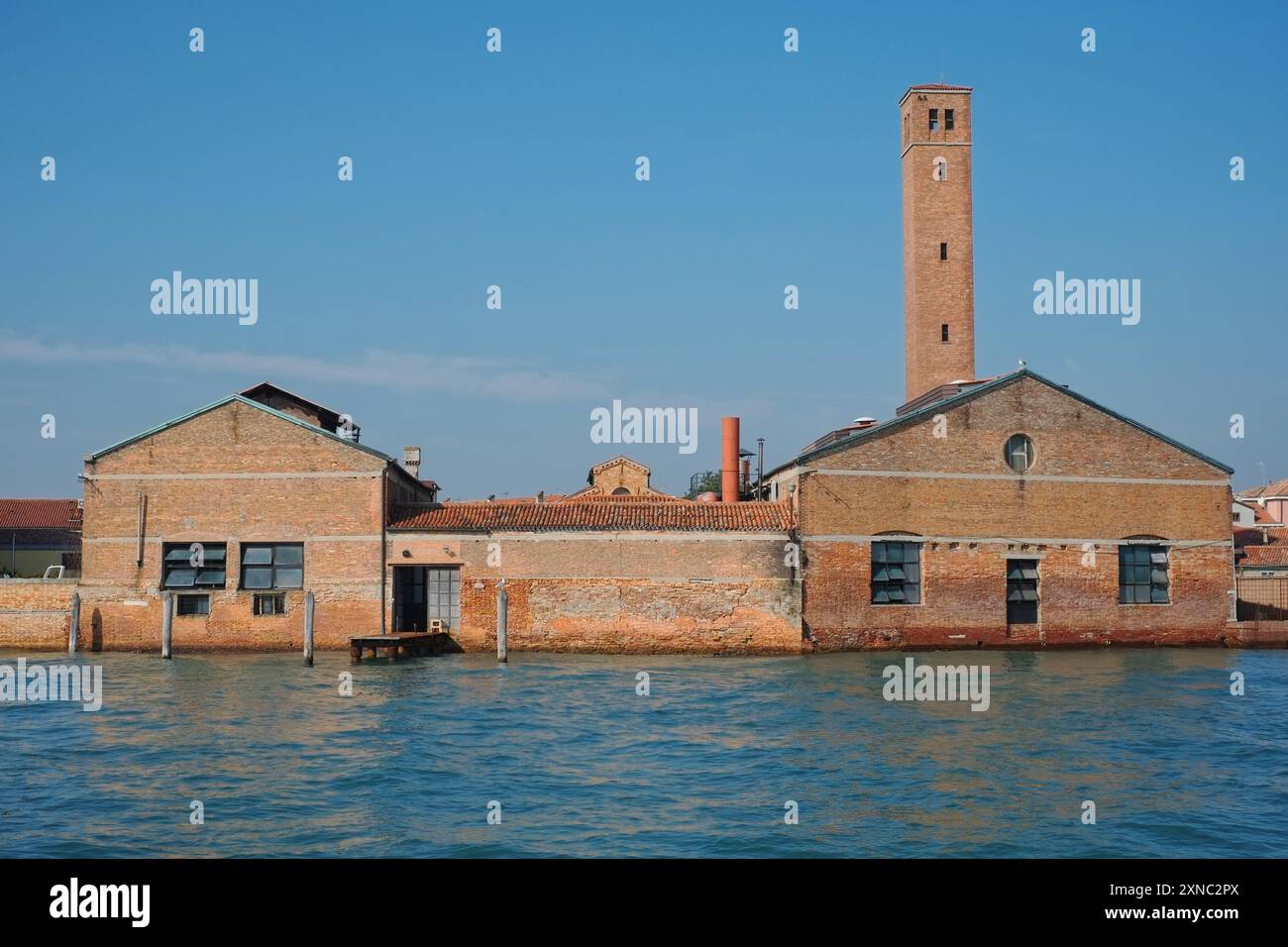 Le mura della fabbrica di Murano di Salviati si affacciano sull'acqua, la punta di una chiesa e un alto campanile inclinabile, il cielo blu, l'acqua turchese a Murano, Venezia Foto Stock