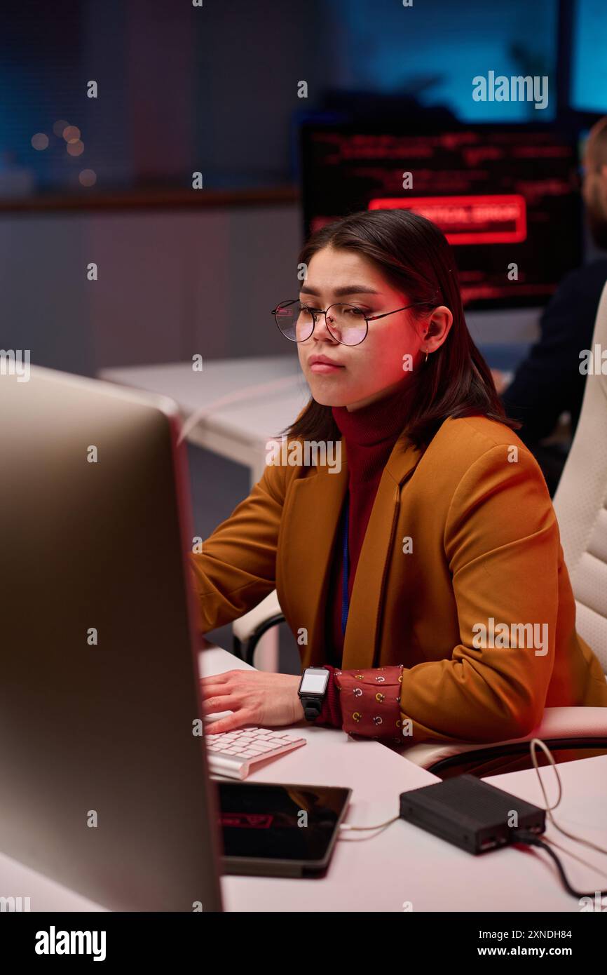Ritratto verticale di una giovane esperta donna che usa il computer nel dipartimento di sicurezza informatica con luci al neon rosse Foto Stock