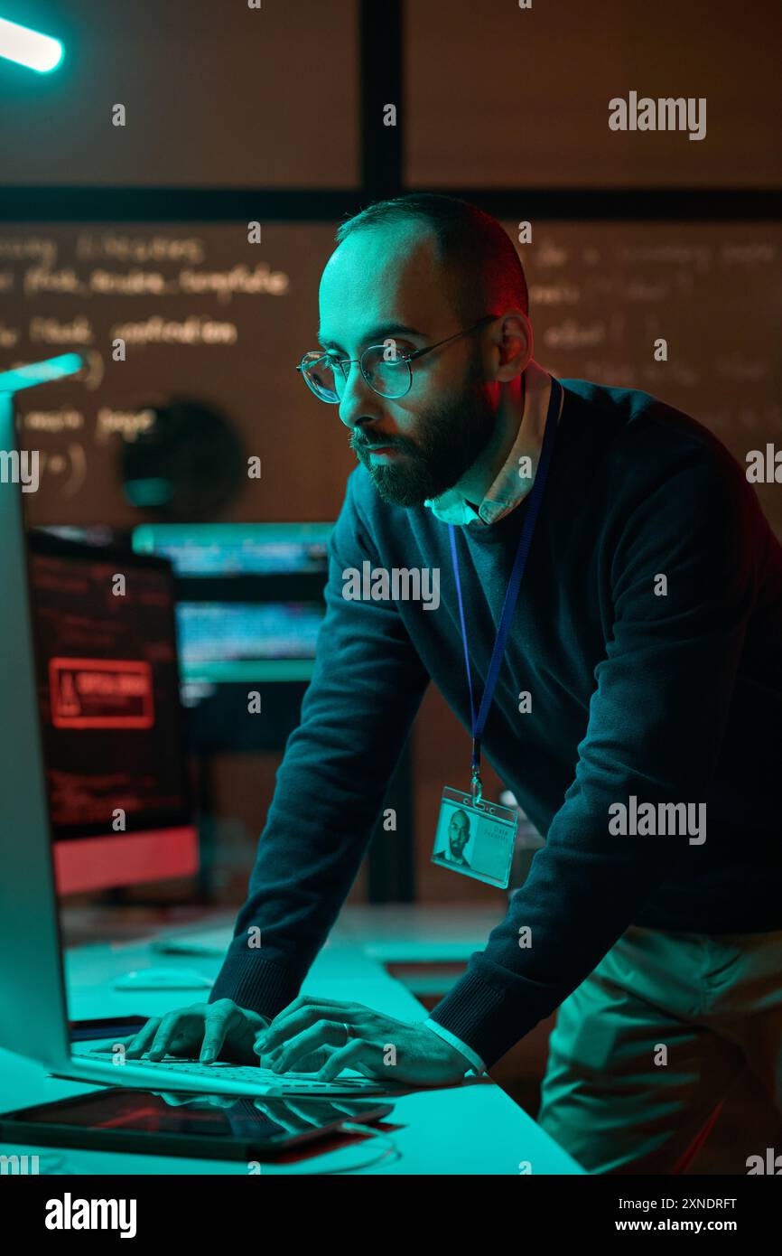 Ritratto verticale di un tecnico barbuto della sicurezza informatica che lavora con una rete di computer con luci al neon blu Foto Stock