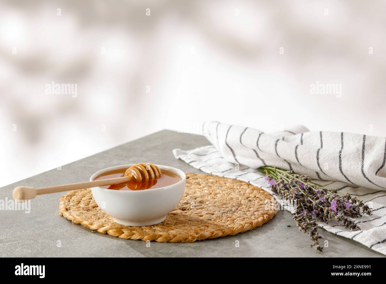 Ciotola bianca di miele e cucchiaia di miele e lavanda su un panno da cucina su un tavolo grigio con sfondo alla luce del sole. Foto Stock