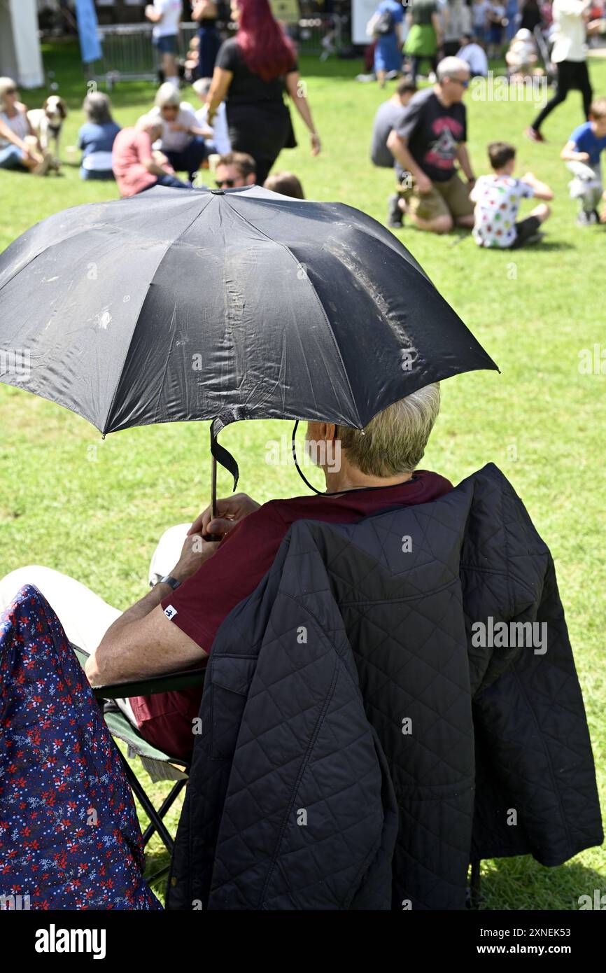 Persona seduta sotto un ombrello per l'ombra al sole al festival all'aperto, famiglie, persone in sottofondo, Regno Unito Foto Stock
