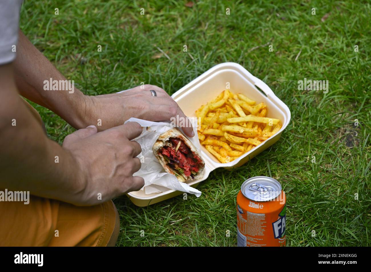 Uomo che mangia il kebab del fast food avvolge il takeaway con patatine e lattina di bibita mentre è seduto sull'erba Foto Stock