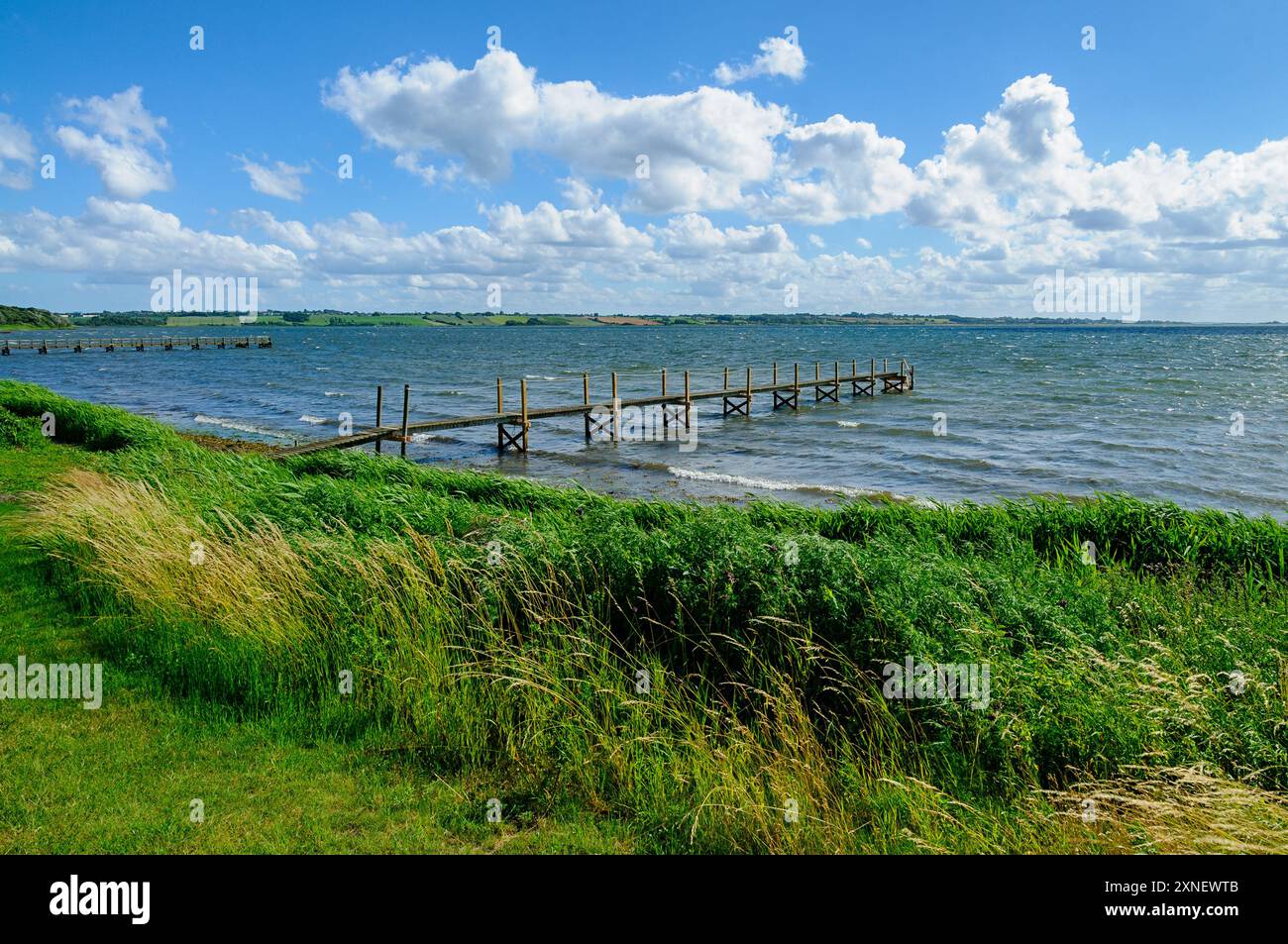 Le dolci onde si infrangono su un molo di legno mentre le soffici nuvole si spostano sopra il lago scintillante circondato da lussureggianti prati verdi. Foto Stock