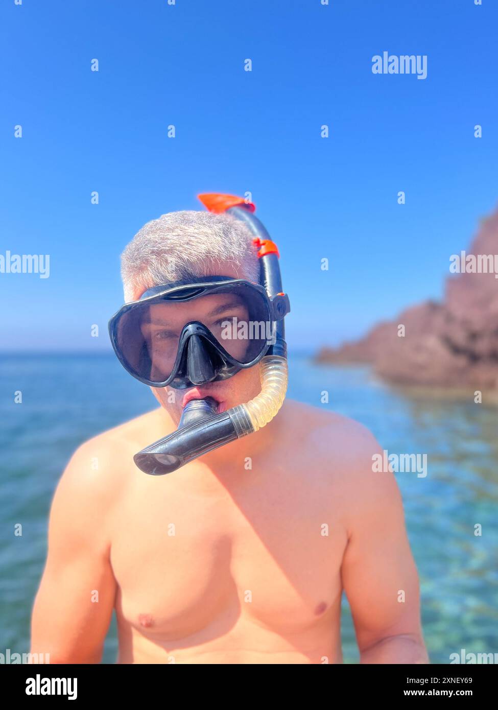 Un uomo sta guardando la macchina fotografica, indossando l'attrezzatura da snorkeling e occhiali di protezione, pronto a tuffarsi in acqua. Lo sfondo mostra cieli azzurri e scogliere rocciose Foto Stock