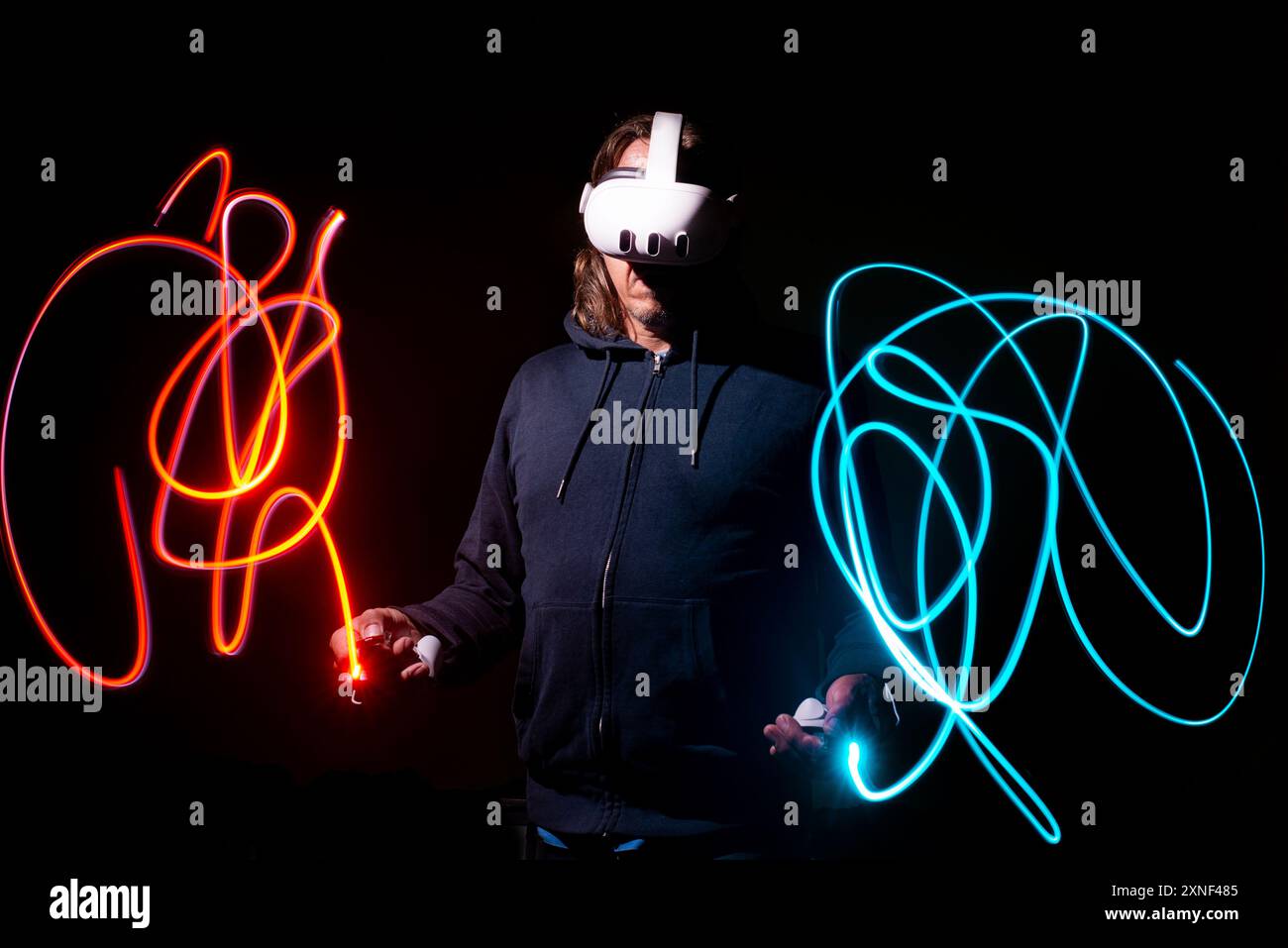 Uomo che indossa un visore Meta quest 3 immerso nella realtà virtuale, circondato da percorsi di luce al neon in un dipinto di luce a lunga esposizione. Foto Stock