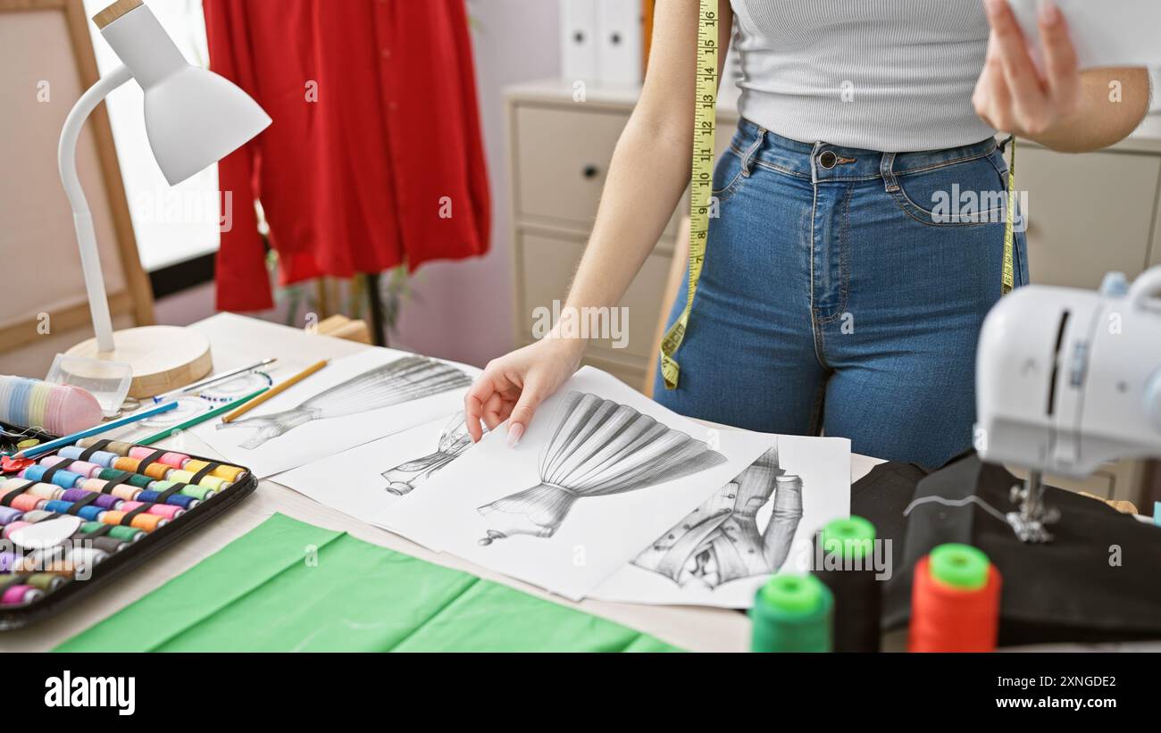 Una donna stilista rivede gli schizzi in un atelier di cucito con tessuti e una macchina da cucire visibile. Foto Stock