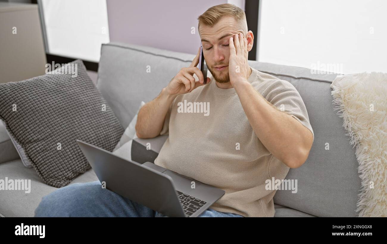 Stressato giovane uomo caucasico con la barba che usa il computer portatile e il telefono negli interni di casa. Foto Stock