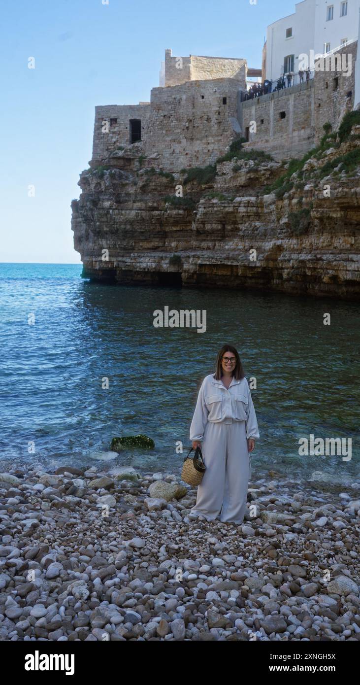 Una giovane donna ispanica si erge sulla spiaggia rocciosa di polignano a mare in puglia, italia, con bellissimi edifici sulla scogliera sullo sfondo e il cl Foto Stock