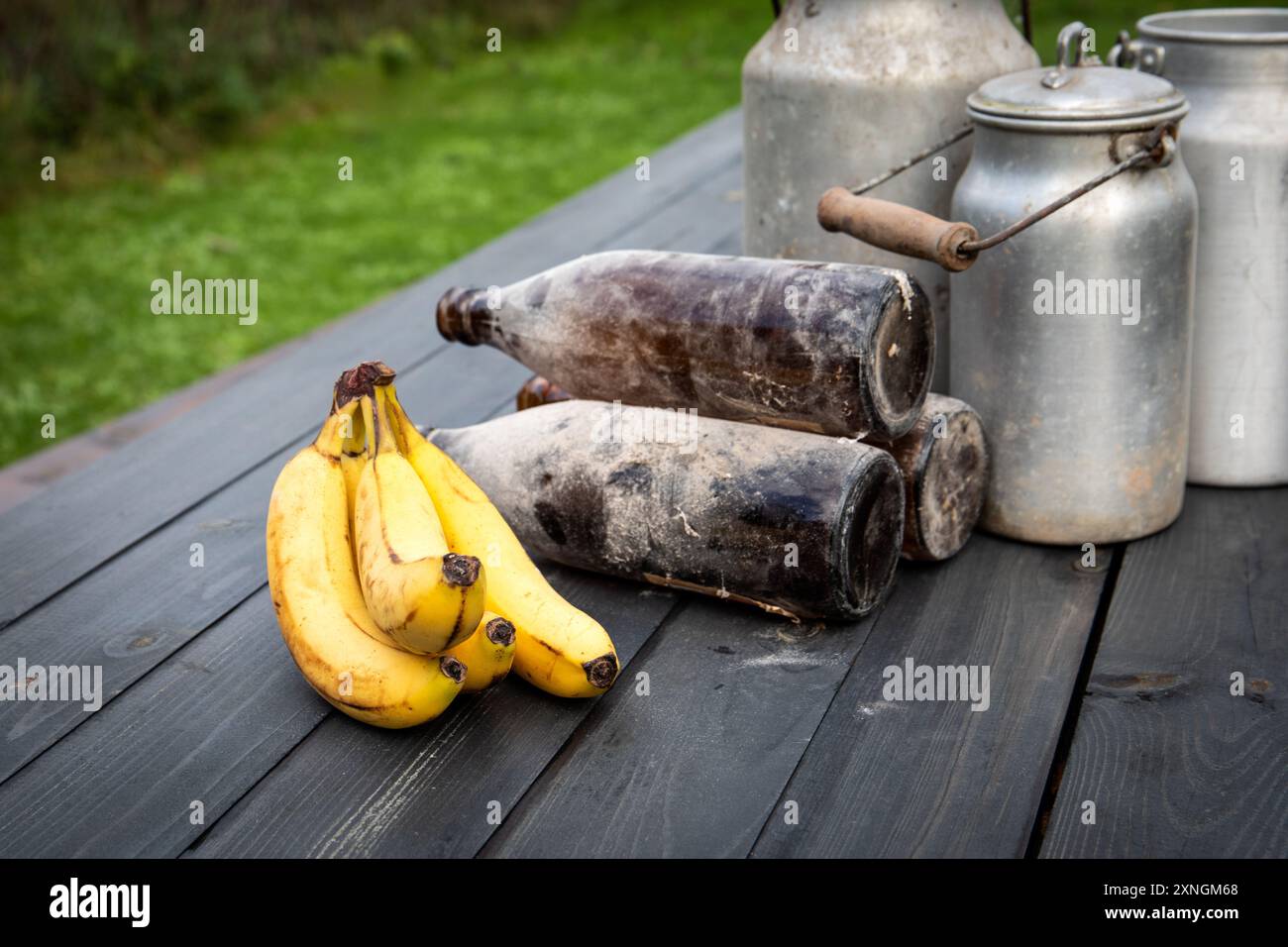 Banane, vecchie bottiglie e lattine di metallo su un tavolo di legno, composizione in un ambiente rurale. Foto Stock