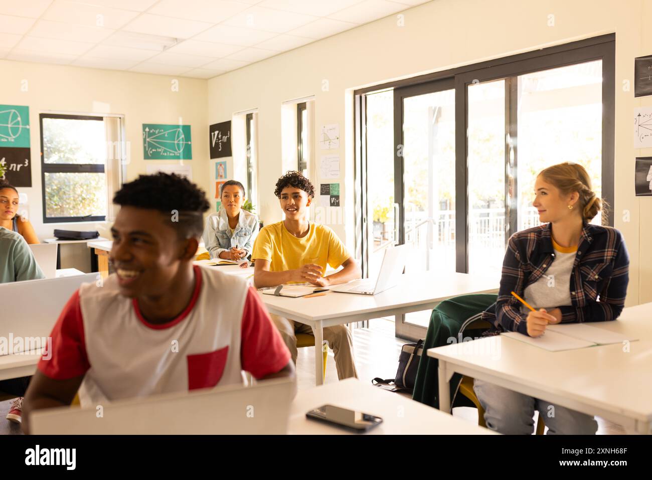 Al liceo, gli adolescenti seduti alla scrivania con il computer portatile, sorridono e si impegnano in classe Foto Stock