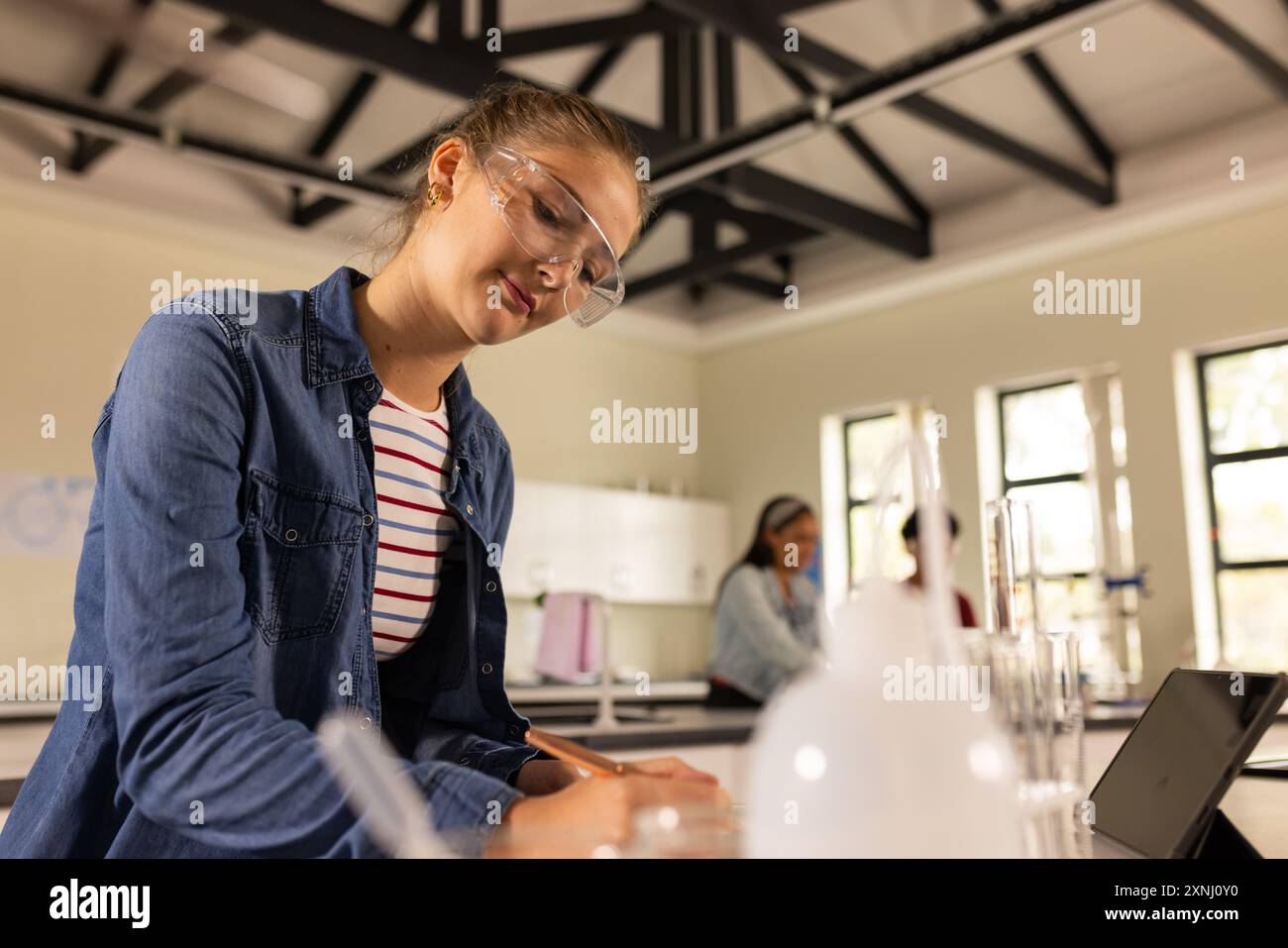 Al liceo, adolescente che scrive appunti in classe scientifica, indossando occhiali di sicurezza Foto Stock