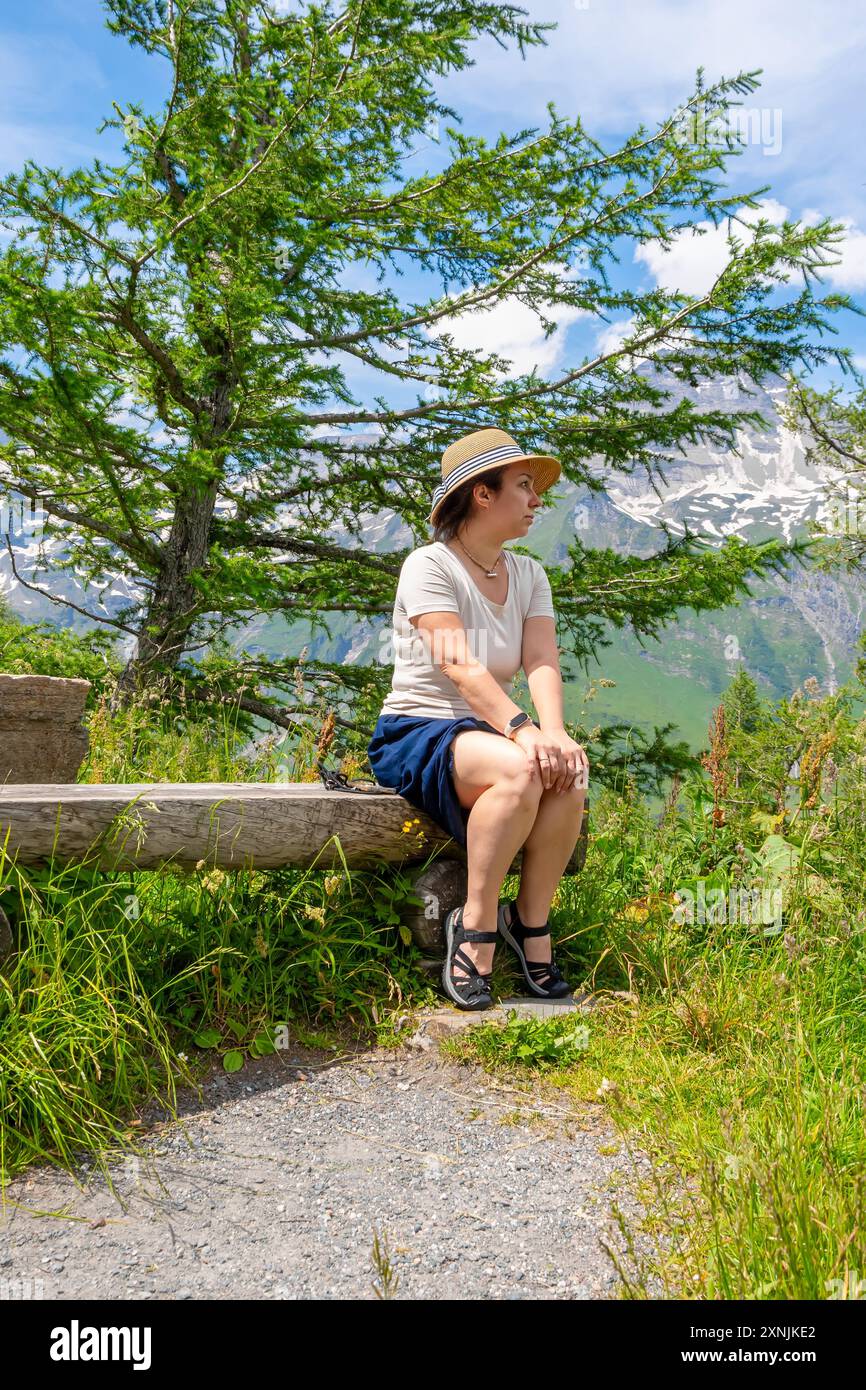 Una donna di 40-45 anni siede circondata da una vegetazione lussureggiante e da una natura colorata. Sullo sfondo, le montagne innevate si innalzano maestosamente sotto il limpido Foto Stock