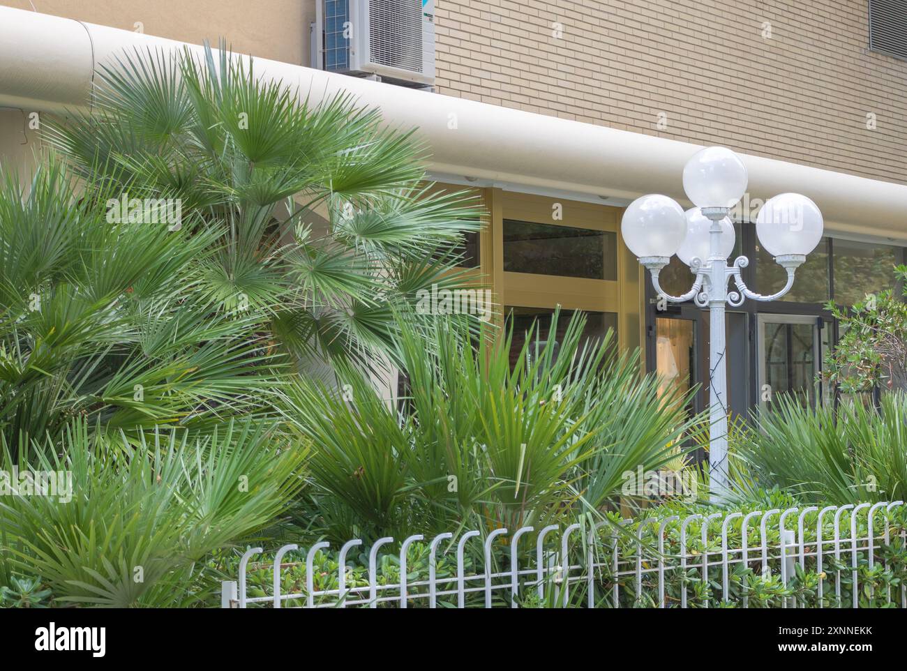 Palme e lampioni bianchi davanti ad un condominio con pareti beige e recinzione bianca. Foto Stock