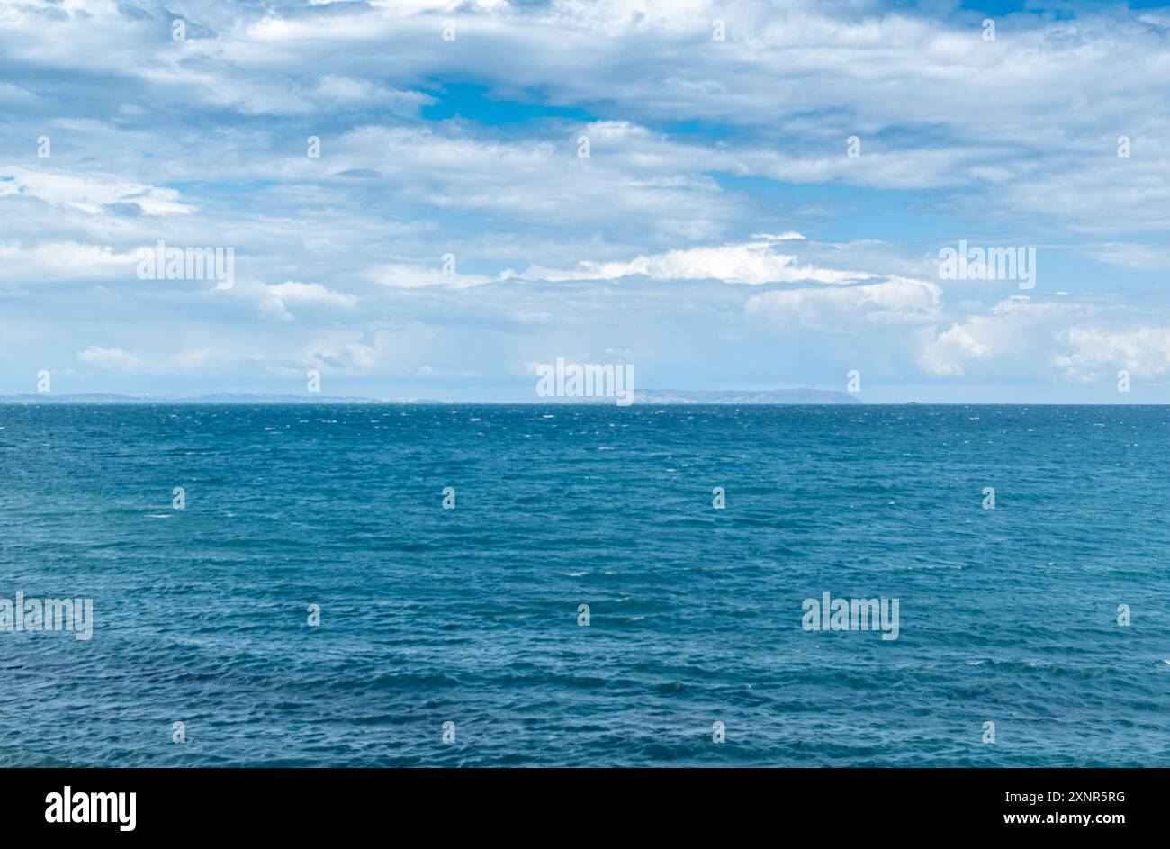 Vista panoramica di un mare calmo e blu sotto un cielo parzialmente nuvoloso, con un molo roccioso che si estende in acqua, perfetto per i viaggi e gli amanti della natura. Foto Stock