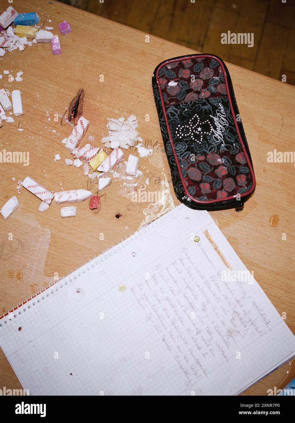 Una scrivania scolastica in disordine con pezzi di gesso rotti, un quaderno lacerato e una custodia a matita, raffigurante le conseguenze di un incidente caotico o violento in una Foto Stock