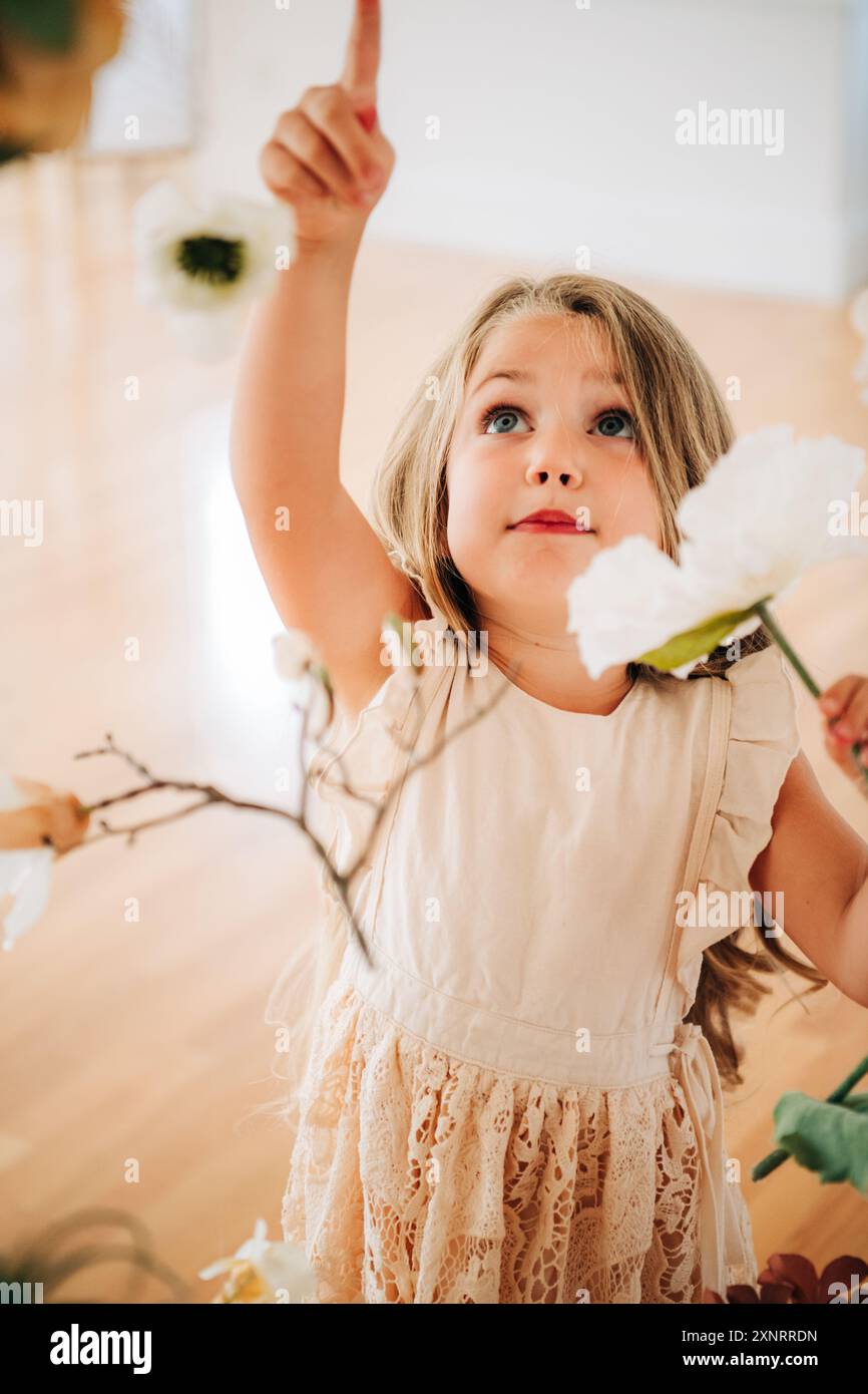 La ragazza bionda con un vestito bianco punta verso l'alto tra i fiori appesi Foto Stock