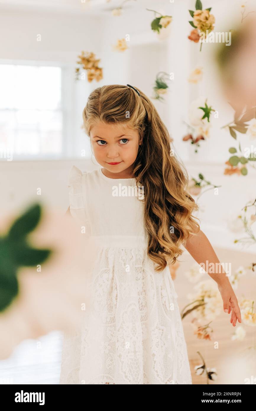 Ragazza bionda in abito bianco tra fiori appesi sembra curiosa Foto Stock
