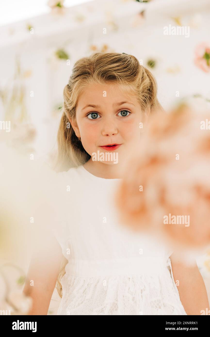 Ragazza bionda in abito bianco tra fiori guarda curiosamente avanti Foto Stock
